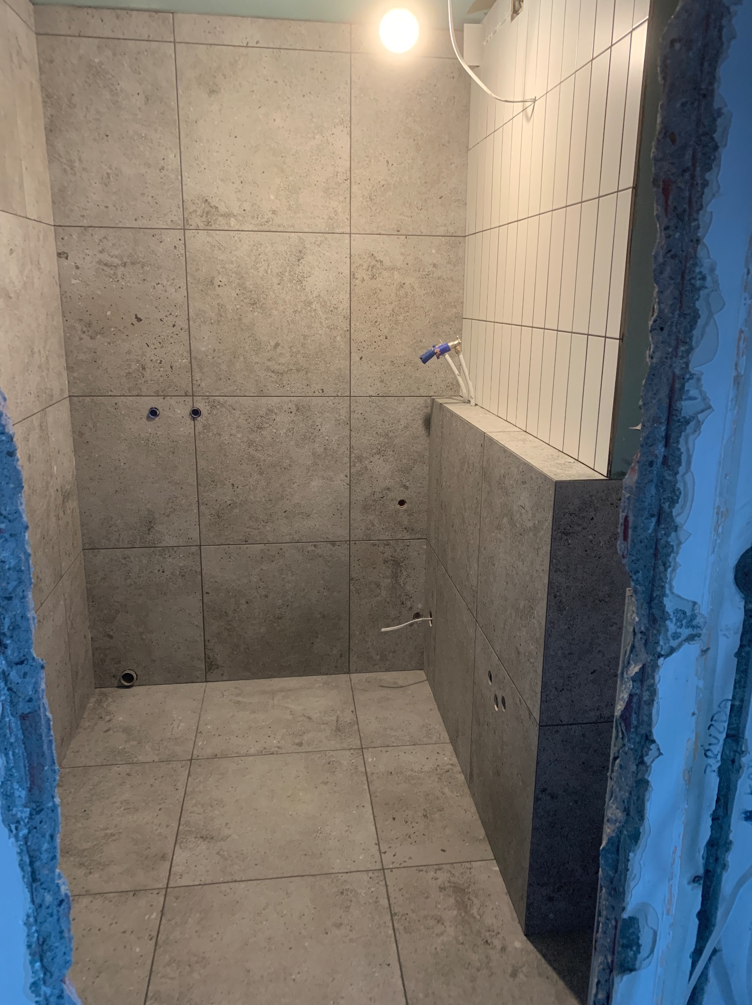 Wnętrze łazienki w trakcie remontu, widoczne szare płytki na ścianach i podłodze, białe płytki na jednej ścianie, wystające rury instalacji wodnej oraz surowy otwór drzwiowy.