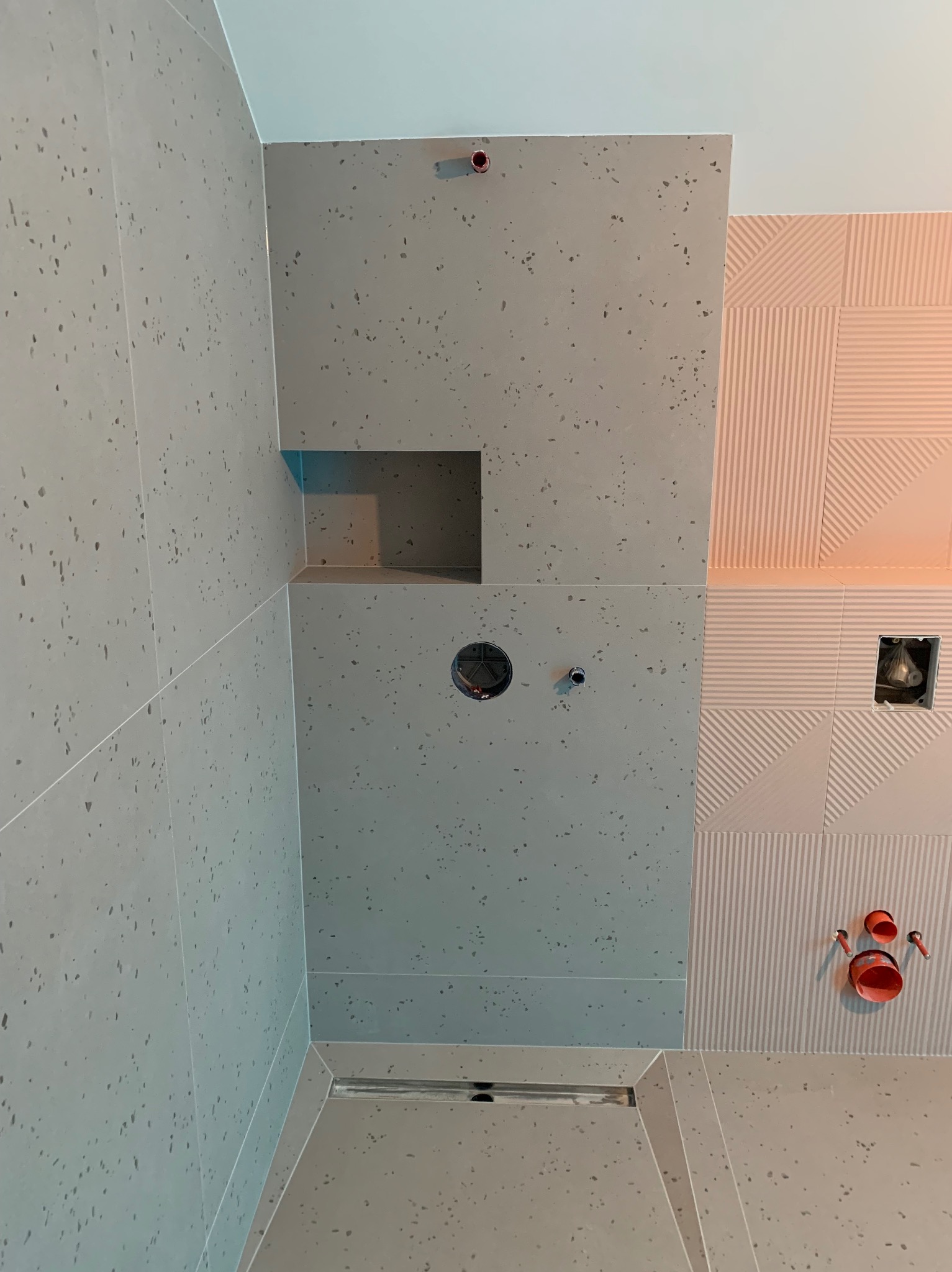 Fragment łazienki w trakcie wykańczania: szare płytki imitujące lastryko, nisza w ścianie, odpływ liniowy w podłodze, instalacja hydrauliczna w ścianie z dekoracyjnymi płytkami w odcieniach różu.