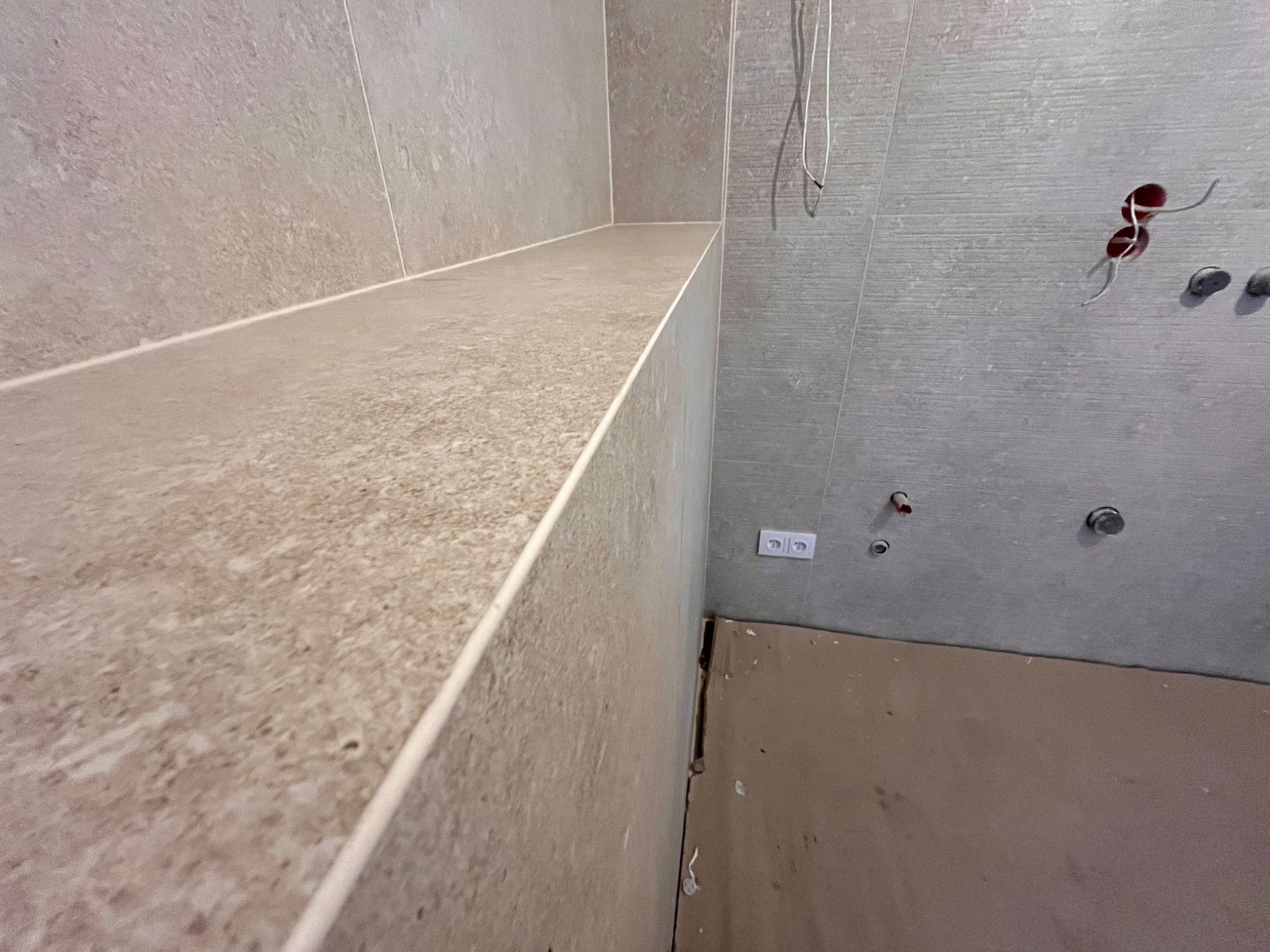 Wykończona półka z płytek w łazience, widoczne gniazdko elektryczne i wystające kable, co sugeruje etap prac wykończeniowych.