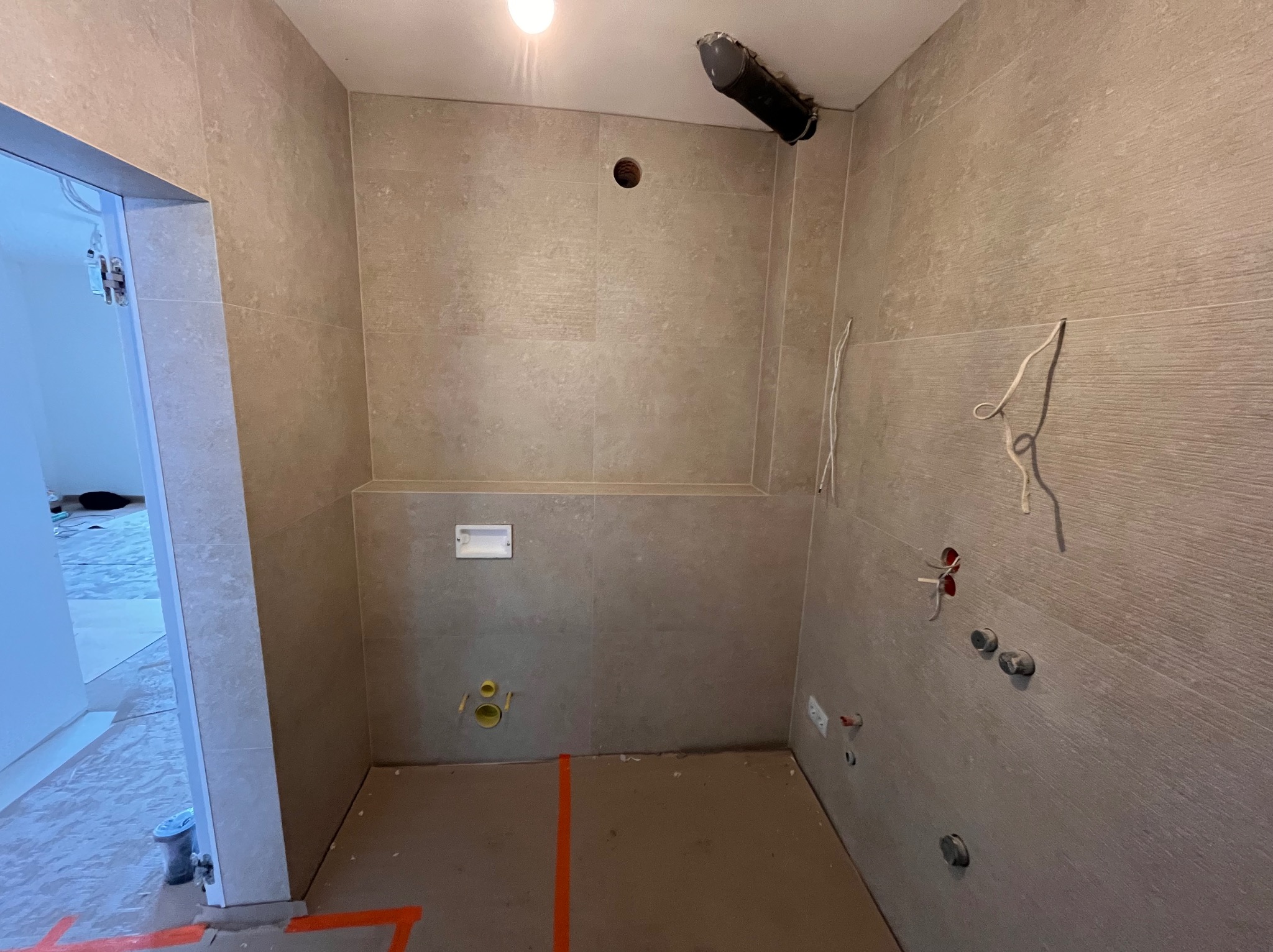 Surowa łazienka w trakcie remontu z beżowymi płytkami, widocznymi instalacjami i wyznaczonymi liniami na podłodze. Otwory na armaturę i oświetlenie.