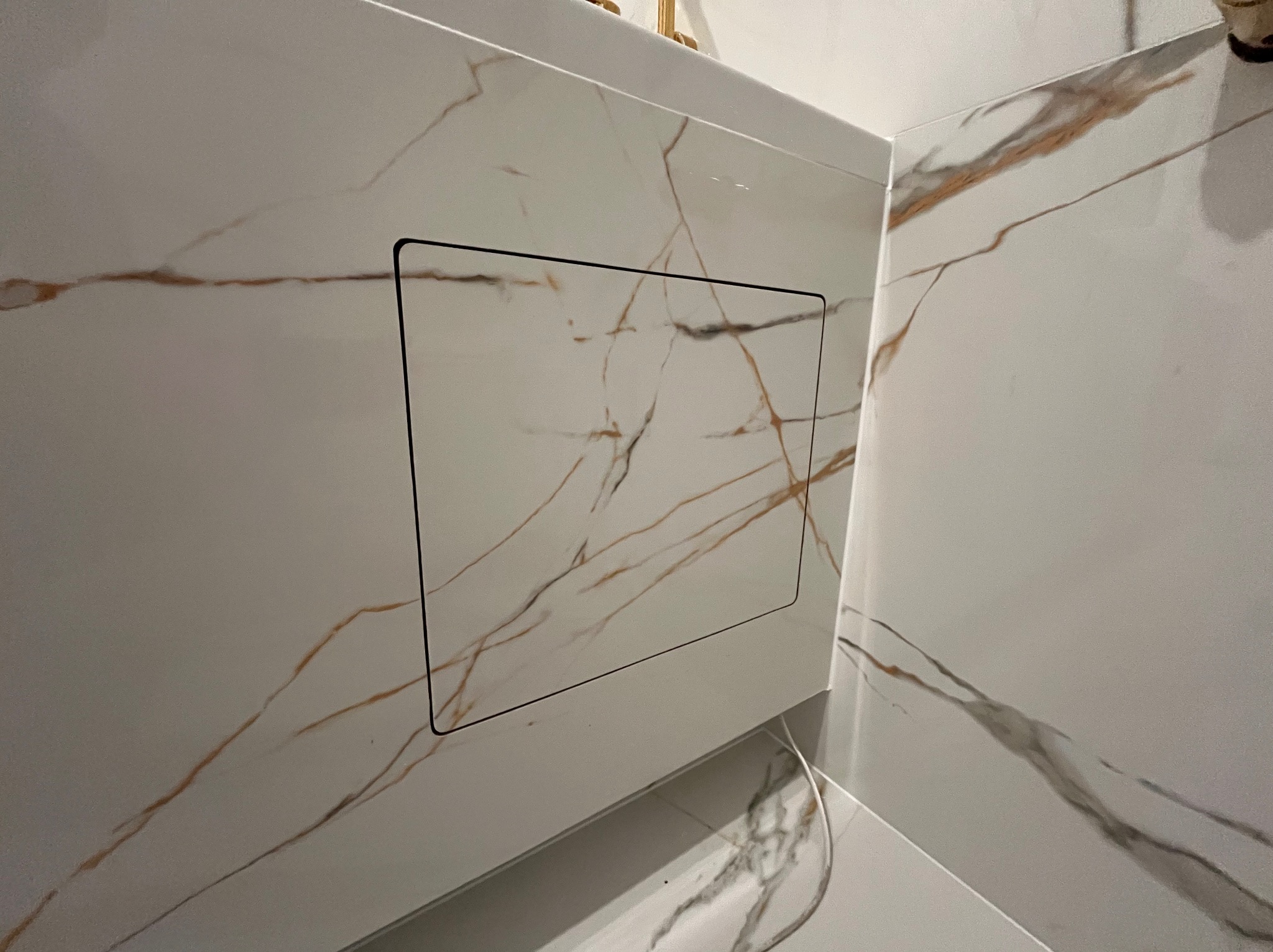 Biała ściana łazienki wyłożona płytkami imitującymi marmur z ukrytym, kwadratowym włazem rewizyjnym z czarną obwódką. Estetyczne wykończenie.