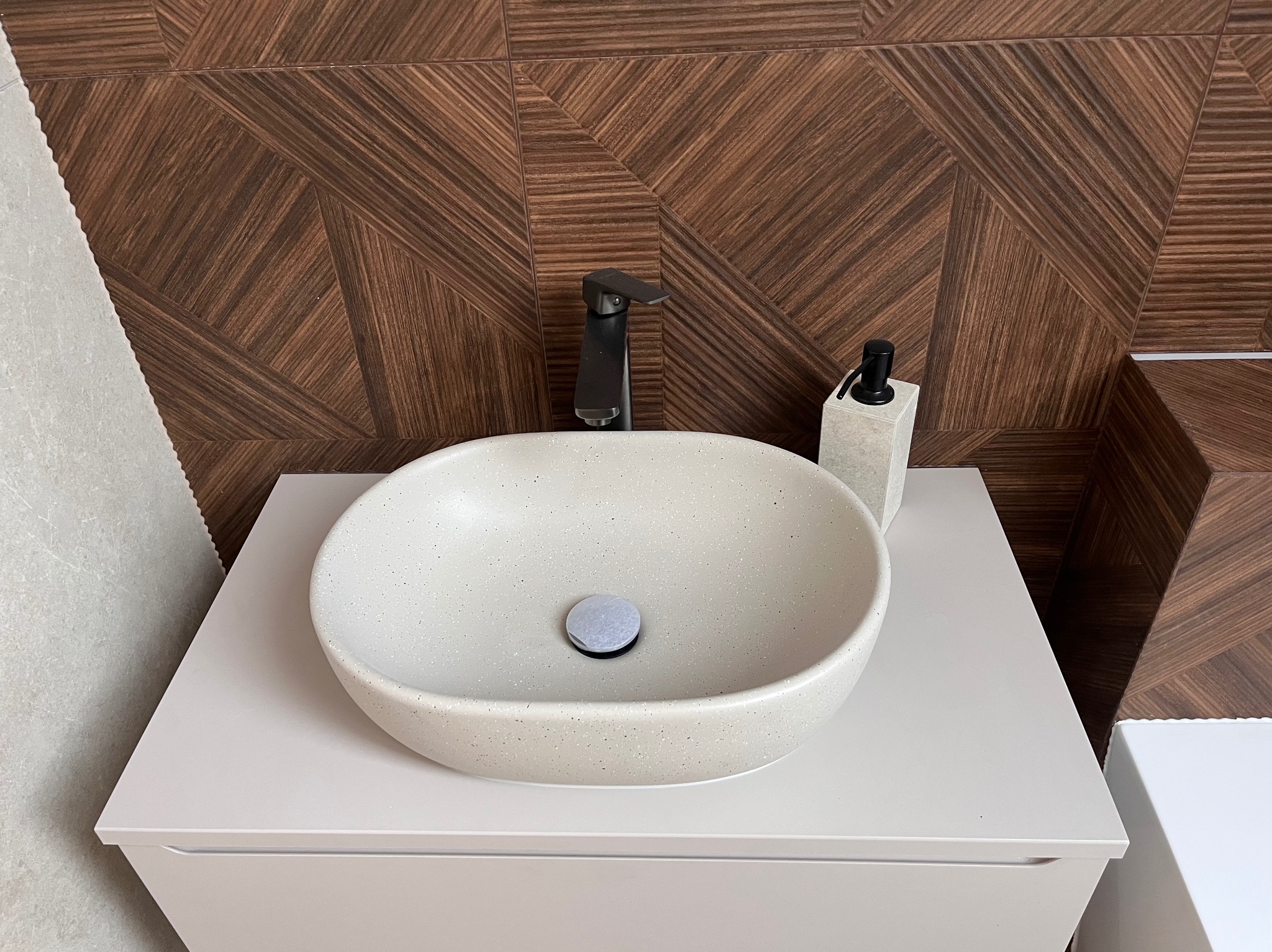Nowoczesna umywalka nablatowa z czarną armaturą i dozownikiem, na tle ściany z płytek imitujących drewno w geometryczny wzór. Minimalistyczny design.
