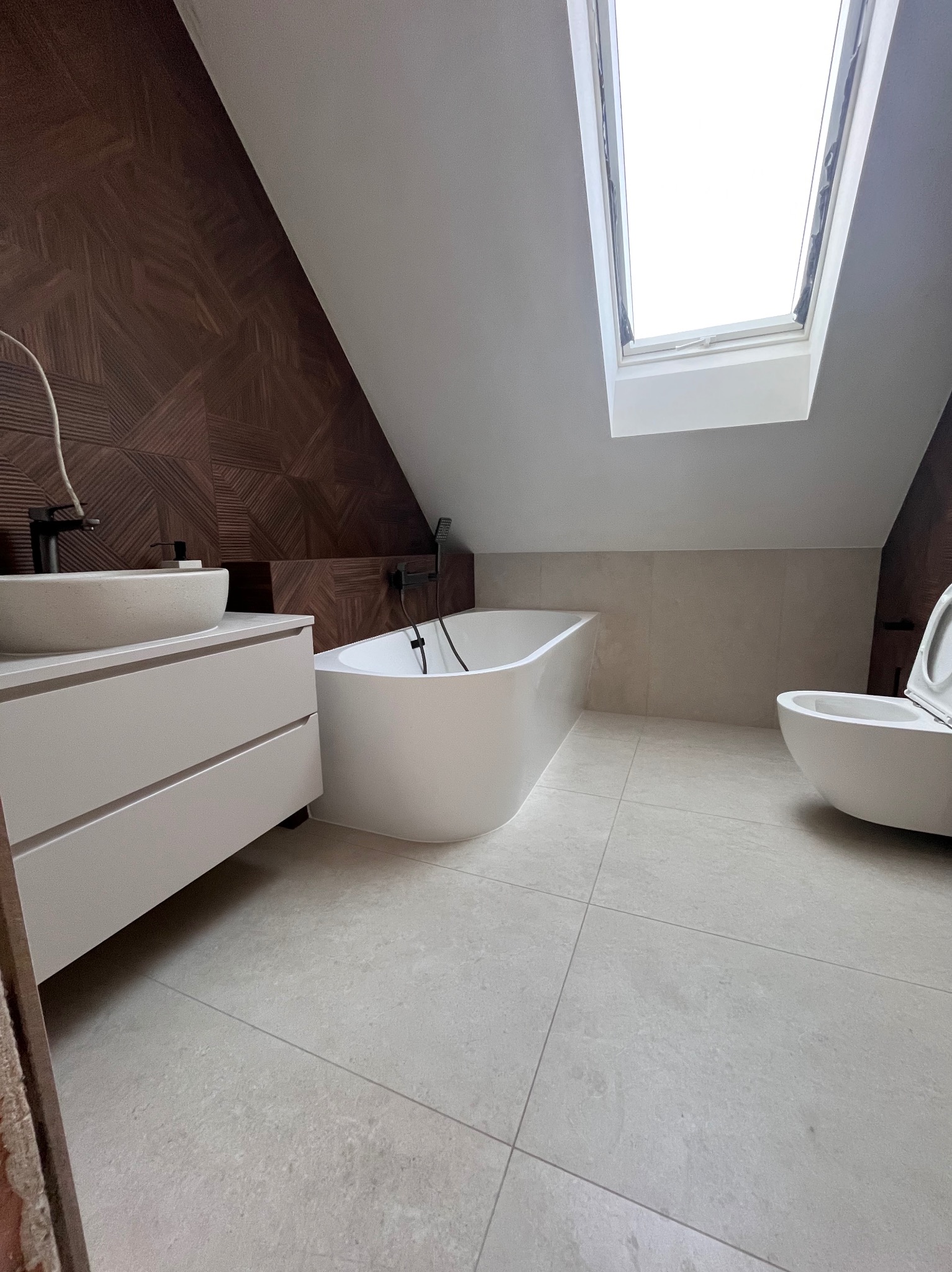 Nowoczesna łazienka na poddaszu z oknem dachowym. Uwagę przykuwa ściana z dekoracyjną, drewnopodobną okładziną. Biała wanna wolnostojąca i umywalka nablatowa.