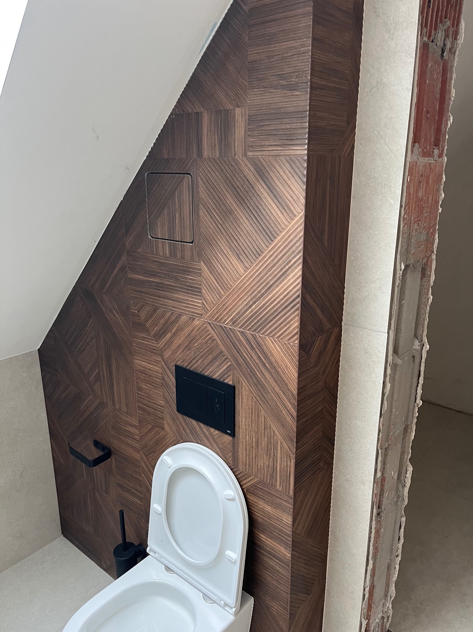 Wykończona toaleta w poddaszu z brązową, wzorzystą okładziną ścienną i białym sanitariatem. Widoczny fragment surowej ściany, podkreślający etap prac.