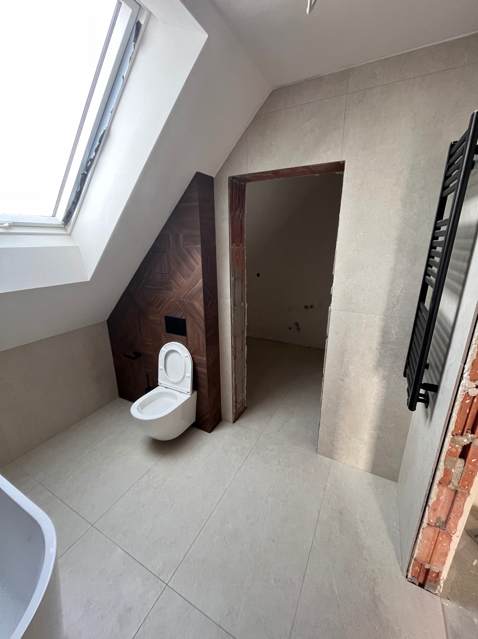 Wykończona łazienka na poddaszu ze skośnym oknem dachowym, z zabudowaną toaletą i grzejnikiem drabinkowym. Widoczne płytki w odcieniach beżu i drewniana okładzina.
