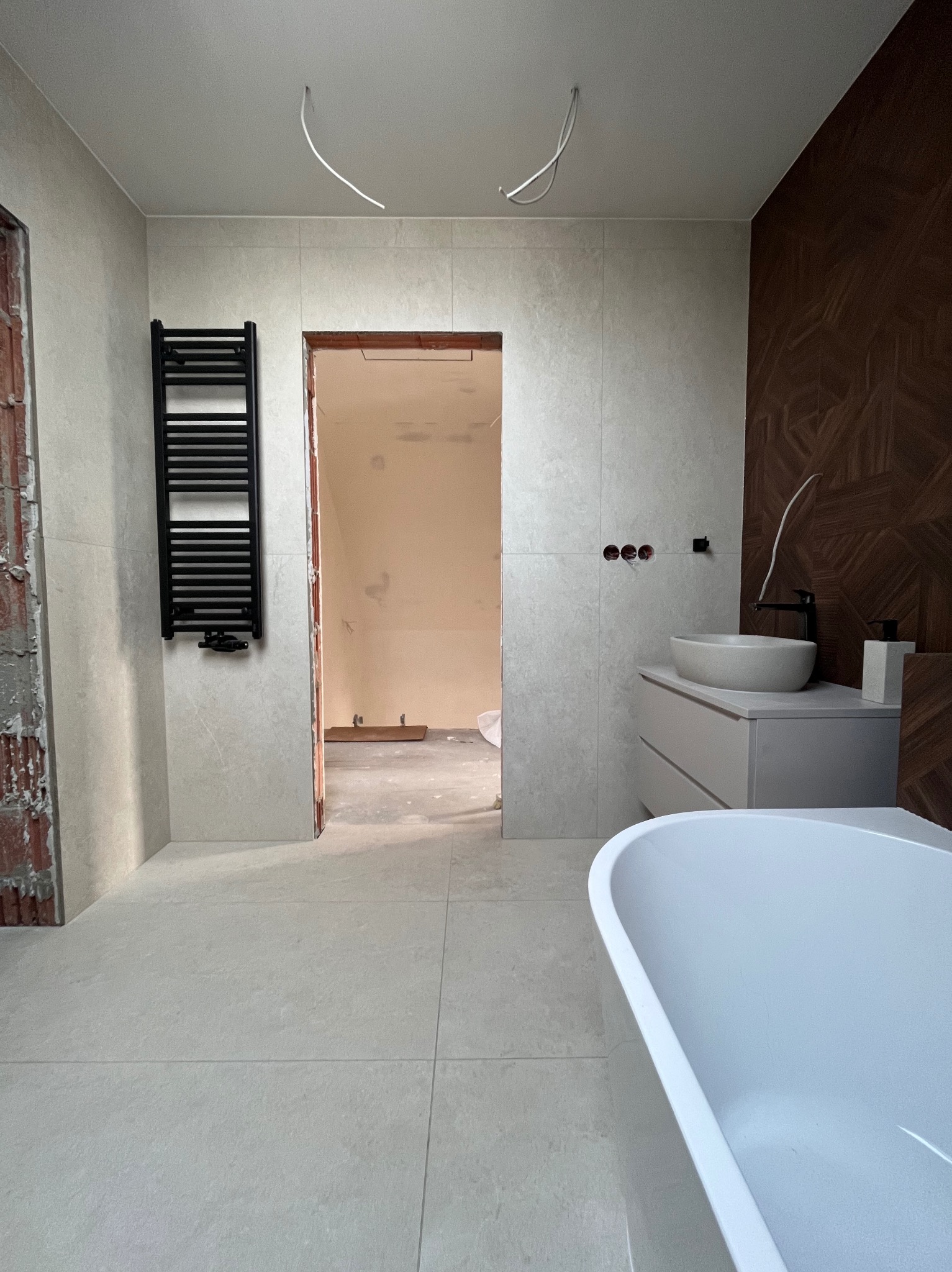 Wnętrze łazienki w trakcie wykańczania: położone płytki, zamontowana umywalka z szafką, grzejnik drabinkowy, widoczne przewody elektryczne. Częściowo widoczna wanna.