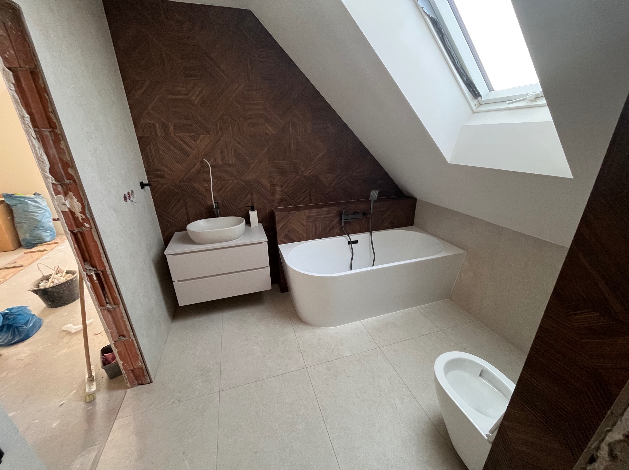 Nowoczesna łazienka na poddaszu z brązową mozaiką na ścianie, białą armaturą i oknem dachowym. Częściowo widoczny stan surowy pomieszczenia.