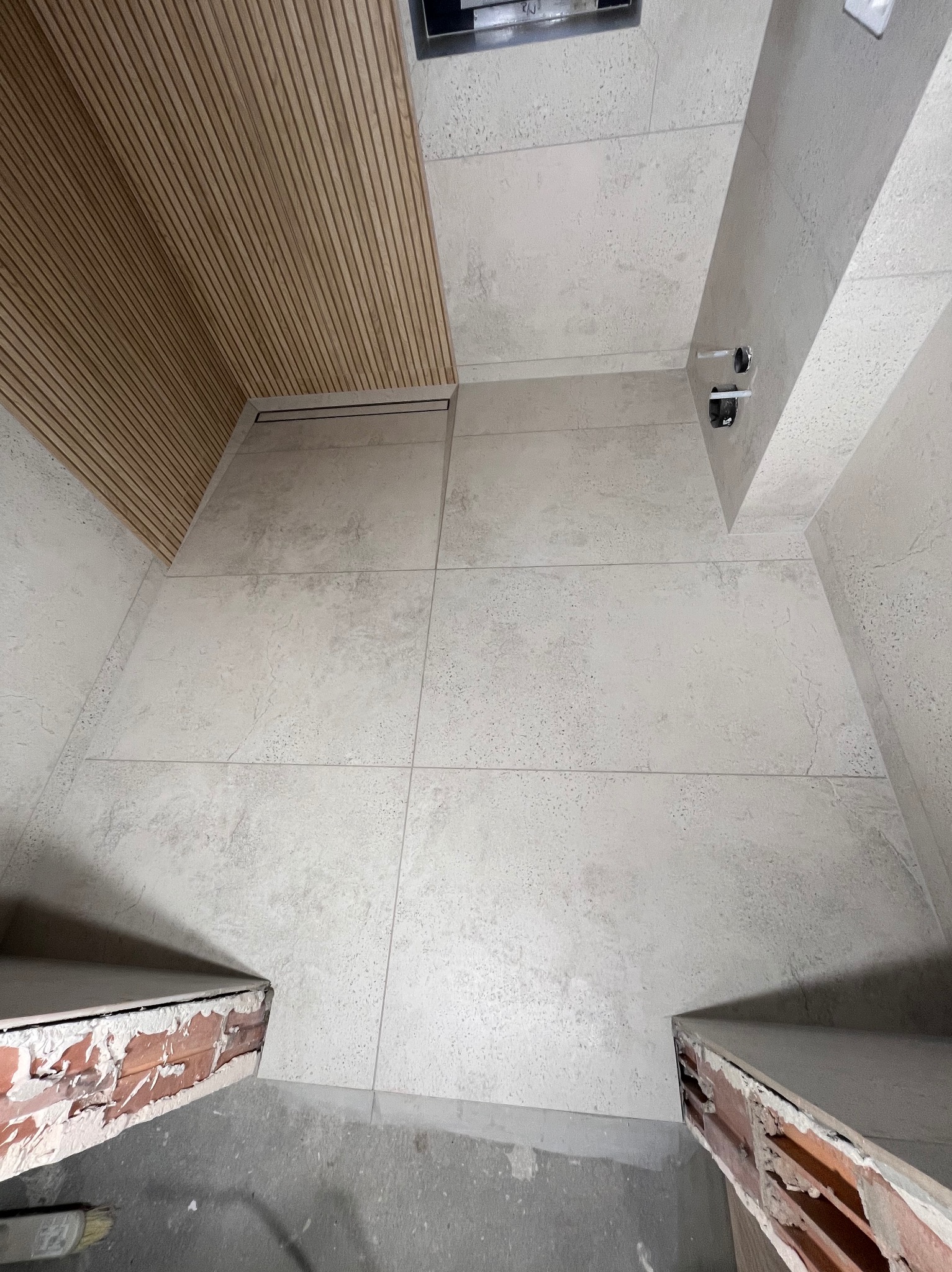 Wykończona łazienka z drewnianym panelem na suficie i jasnymi, dużymi płytkami na ścianach i podłodze. Widoczne przygotowane punkty instalacji wodnej.