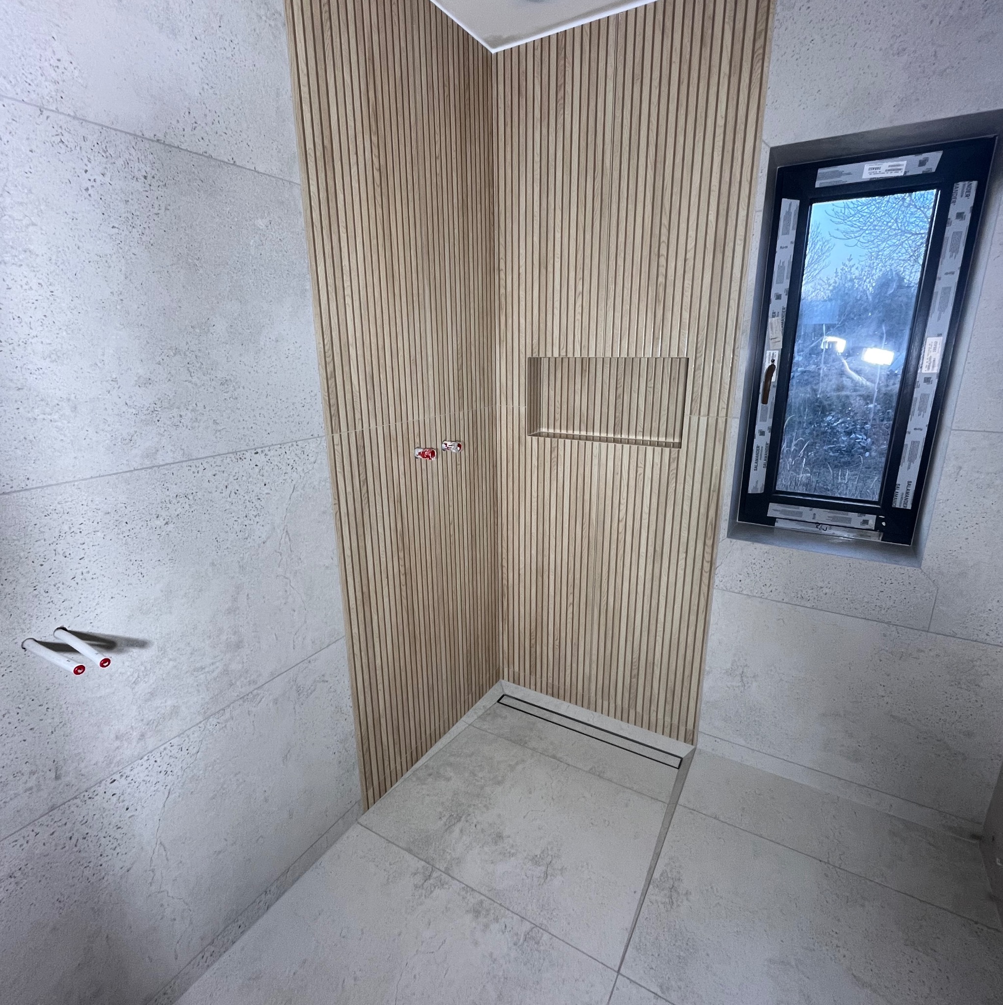 Nowoczesna łazienka z drewnianą ścianą, wnęką i odpływem liniowym. Jasne płytki imitujące kamień na ścianach i podłodze. Okno z czarną ramą.