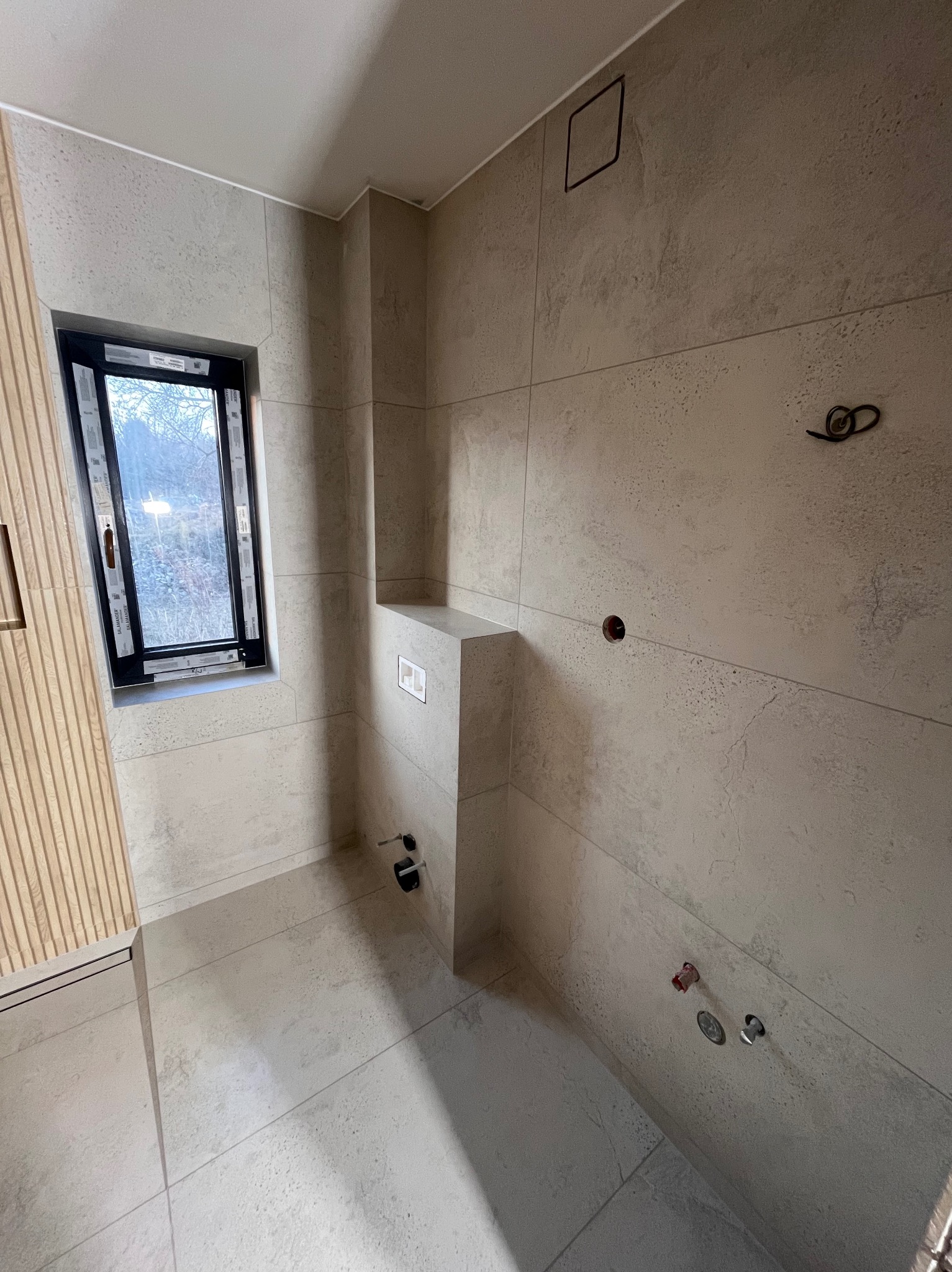 Wykończona łazienka z beżowymi płytkami, widoczne przyłącza wodne i okno z czarną ramą. Minimalistyczny design, surowy stan przed montażem armatury.