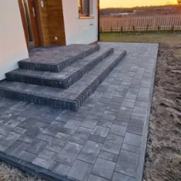 Ułożona szara kostka brukowa tworząca taras i schody wejściowe do domu. W tle widoczny zachód słońca i ogrodzenie posesji.