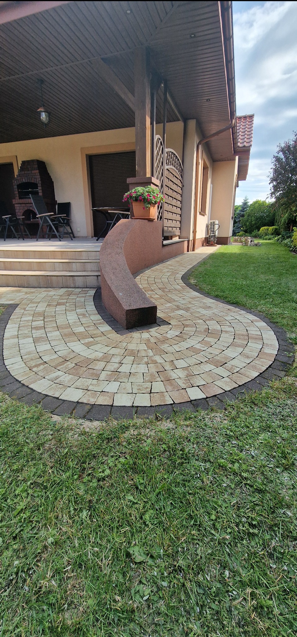 Okrągła ścieżka z kostki brukowej w odcieniach beżu i brązu, prowadząca od domu z kolumnami i kwiatami w donicy, otoczona trawnikiem. Nietypowy wzór ułożenia.