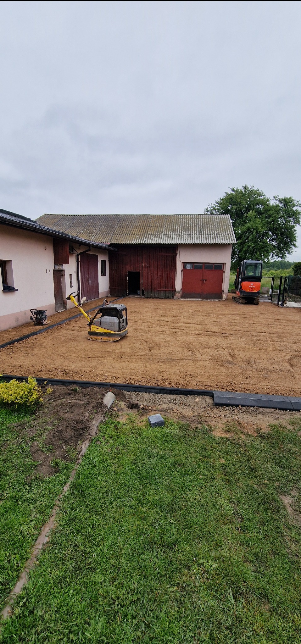 Przygotowany teren pod kostkę brukową z obrzeżami, zagęszczarką i minikoparką na posesji z budynkiem gospodarczym w tle.