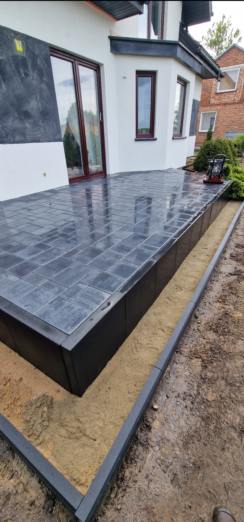 Nowoczesny taras z szarej kostki brukowej, obrzeża w kolorze antracytu. Widoczna ubijarka wibracyjna. Tło stanowi biały budynek z brązowymi oknami.
