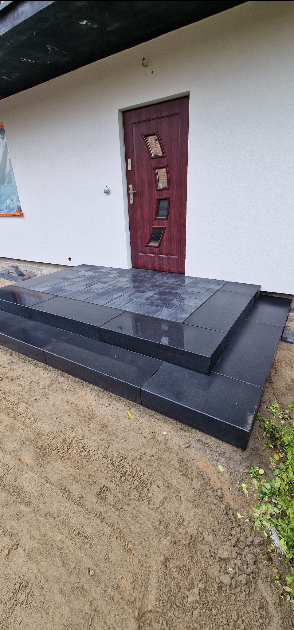 Wykończone schody wejściowe z ciemnej kostki brukowej, prowadzące do brązowych drzwi wejściowych w białym budynku. Kostka ułożona na dwóch stopniach i podest przed drzwiami.