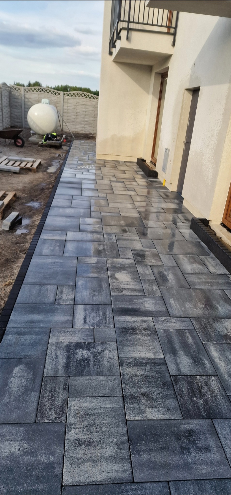 Świeżo ułożona, szara kostka brukowa przed domem z białymi ścianami. Widoczny fragment ogrodzenia i zbiornik na gaz. Ułożona w nieregularny wzór.