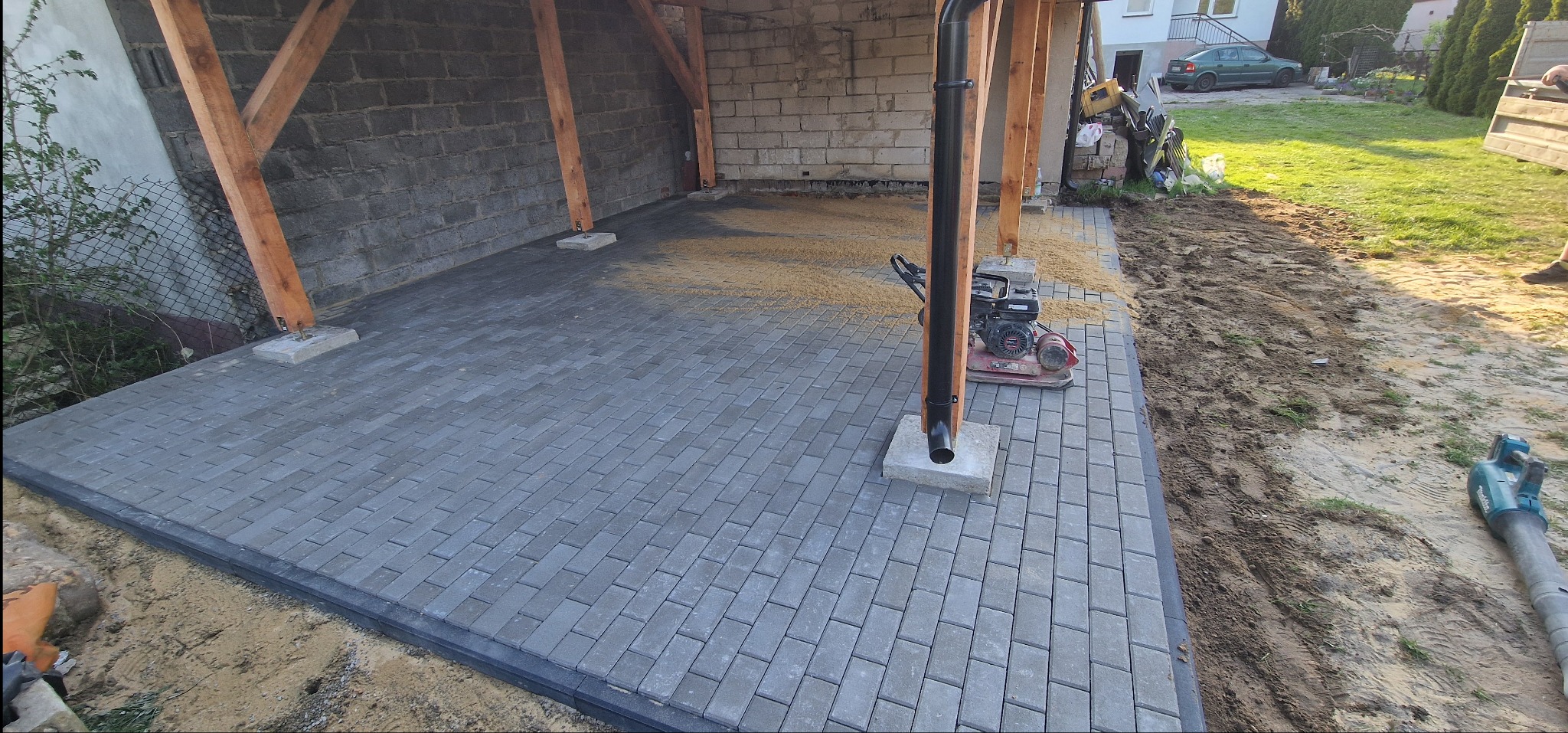 Ułożona szara kostka brukowa pod zadaszeniem, widoczna ubijarka i piasek. Częściowo widoczny teren przygotowany pod dalsze prace brukarskie.