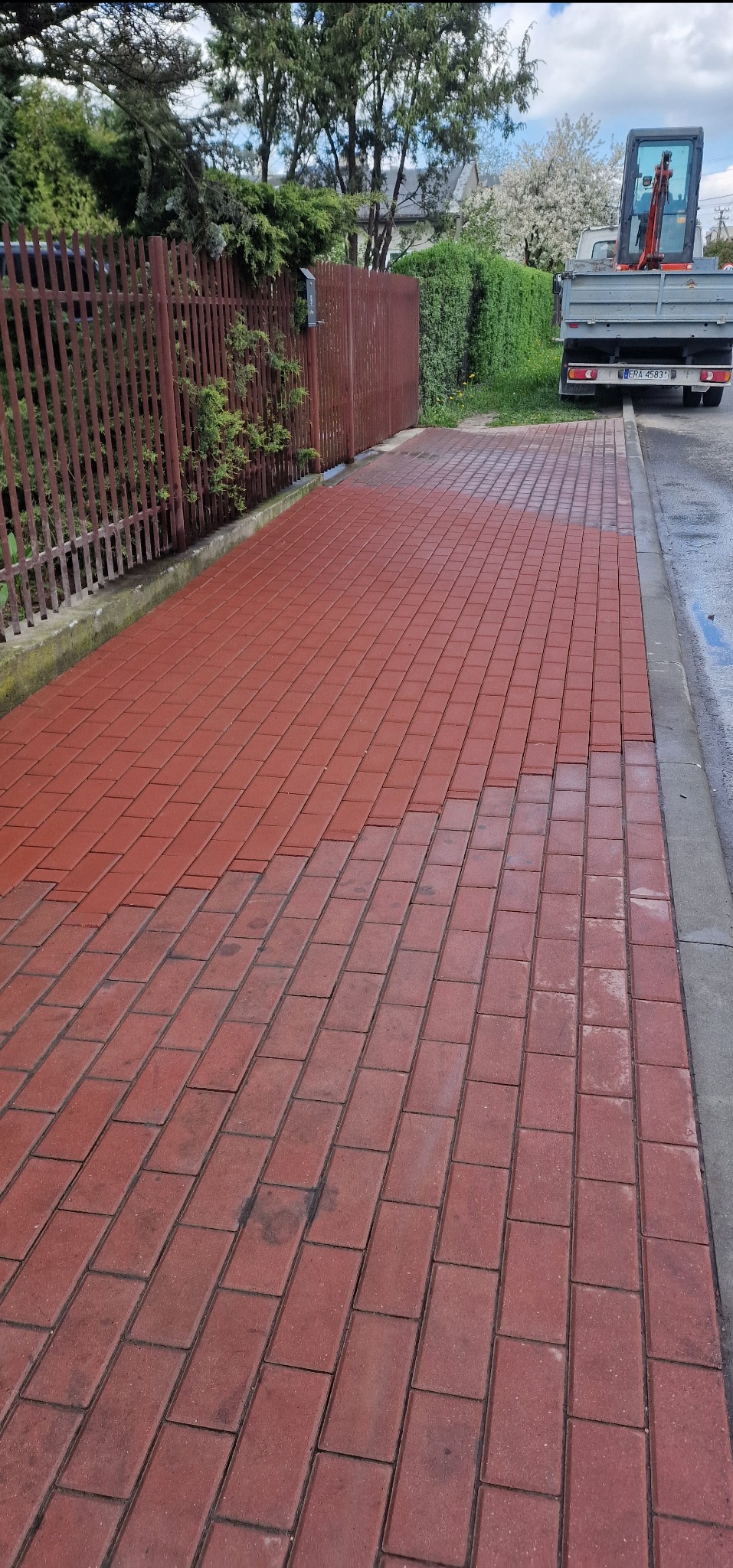 Czerwona kostka brukowa ułożona przy płocie, obok zaparkowany samochód z mini koparką na pace. Część kostki jest mokra, co sugeruje mycie lub świeże ułożenie.