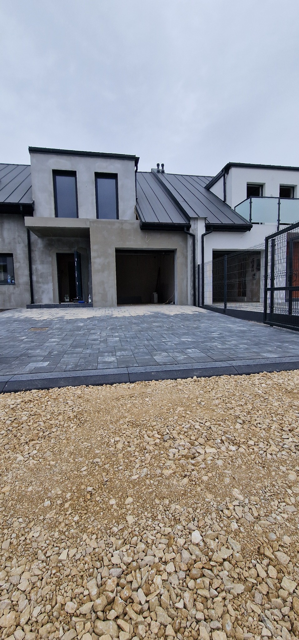 Nowoczesny dom z szarą kostką brukową na podjeździe i podsypką z kruszywa. Widoczny fragment ogrodzenia i surowy stan elewacji budynku.