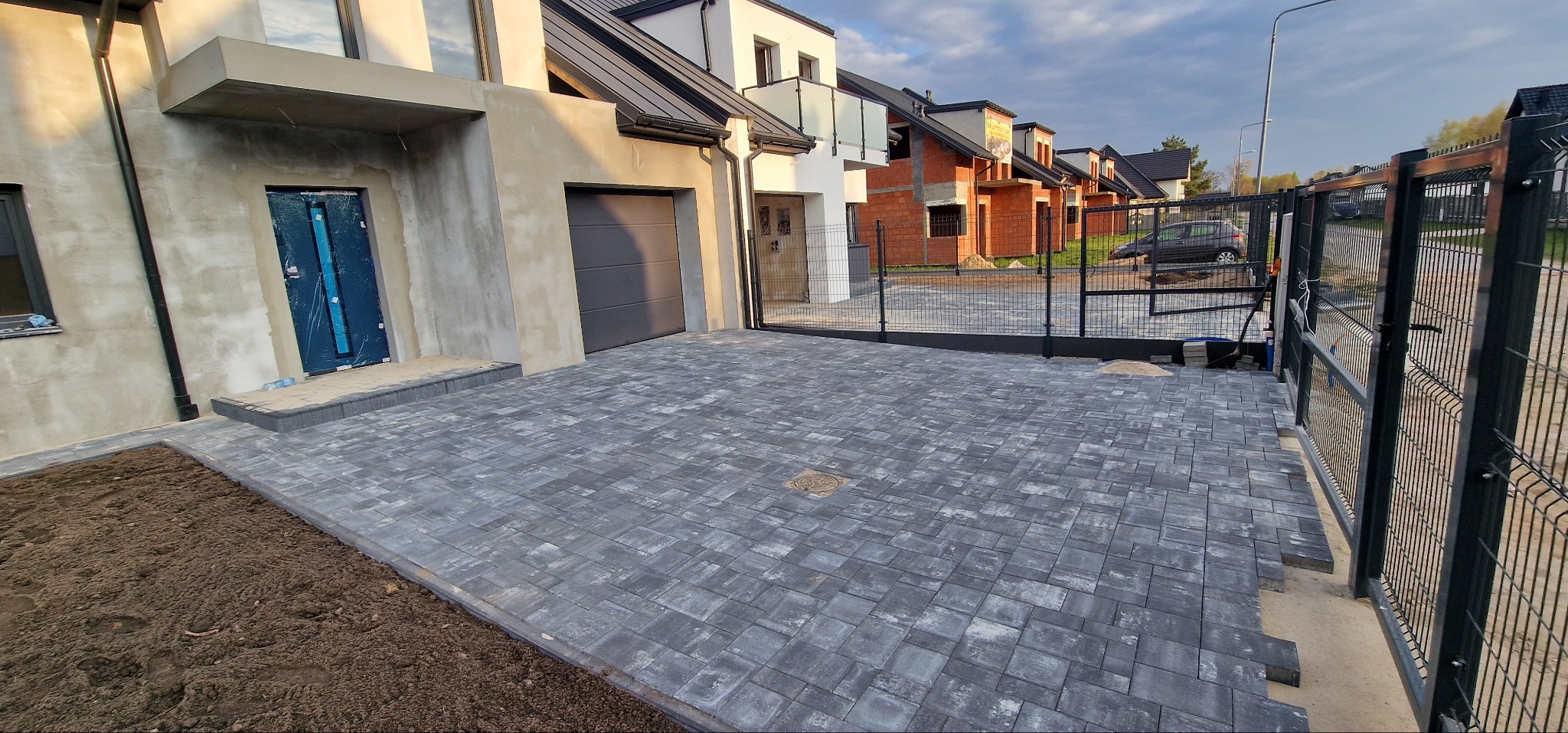 Ułożona kostka brukowa na podjeździe do domu z garażem i furtką. Szara kostka, częściowo odsłonięta ziemia. Nowe budynki w tle, błękitne niebo.