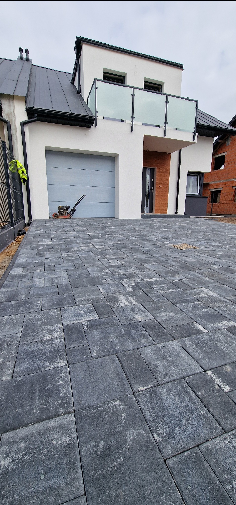 Ułożona kostka brukowa w odcieniach szarości przed nowoczesnym domem z garażem i balkonem. W tle zagęszczarka wibracyjna.