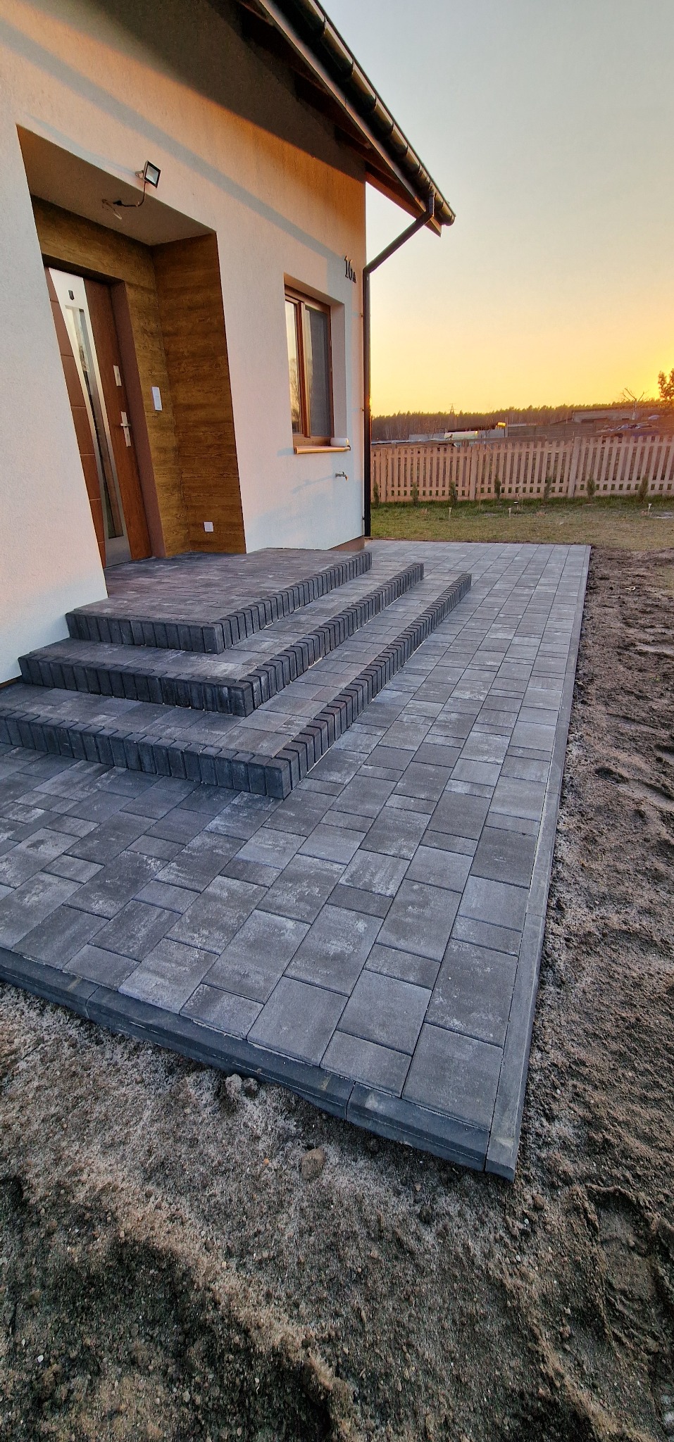 Ułożona szara kostka brukowa tworząca taras i schody wejściowe do domu. W tle widoczny zachód słońca i ogrodzenie posesji.