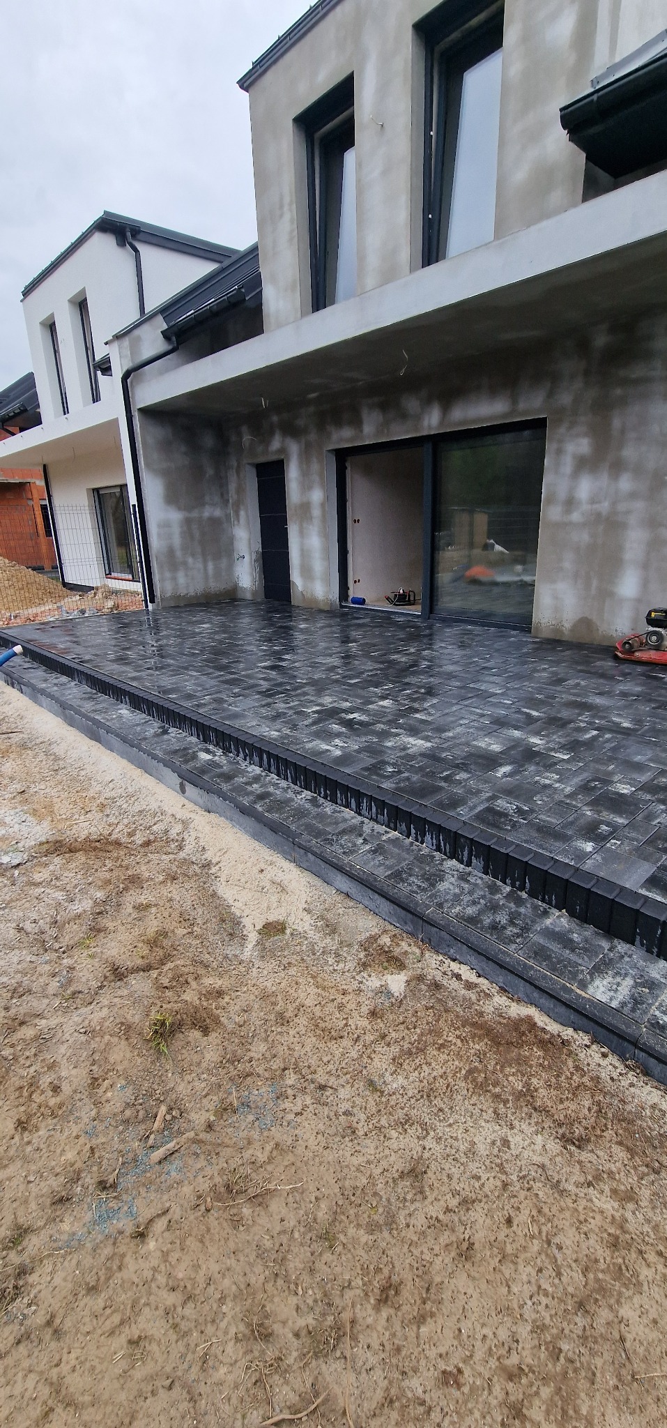 Nowoczesny taras z ciemnej kostki brukowej przy domu w trakcie wykańczania. Widoczny fragment gruntu i sprzęt budowlany. Surowy beton elewacji.