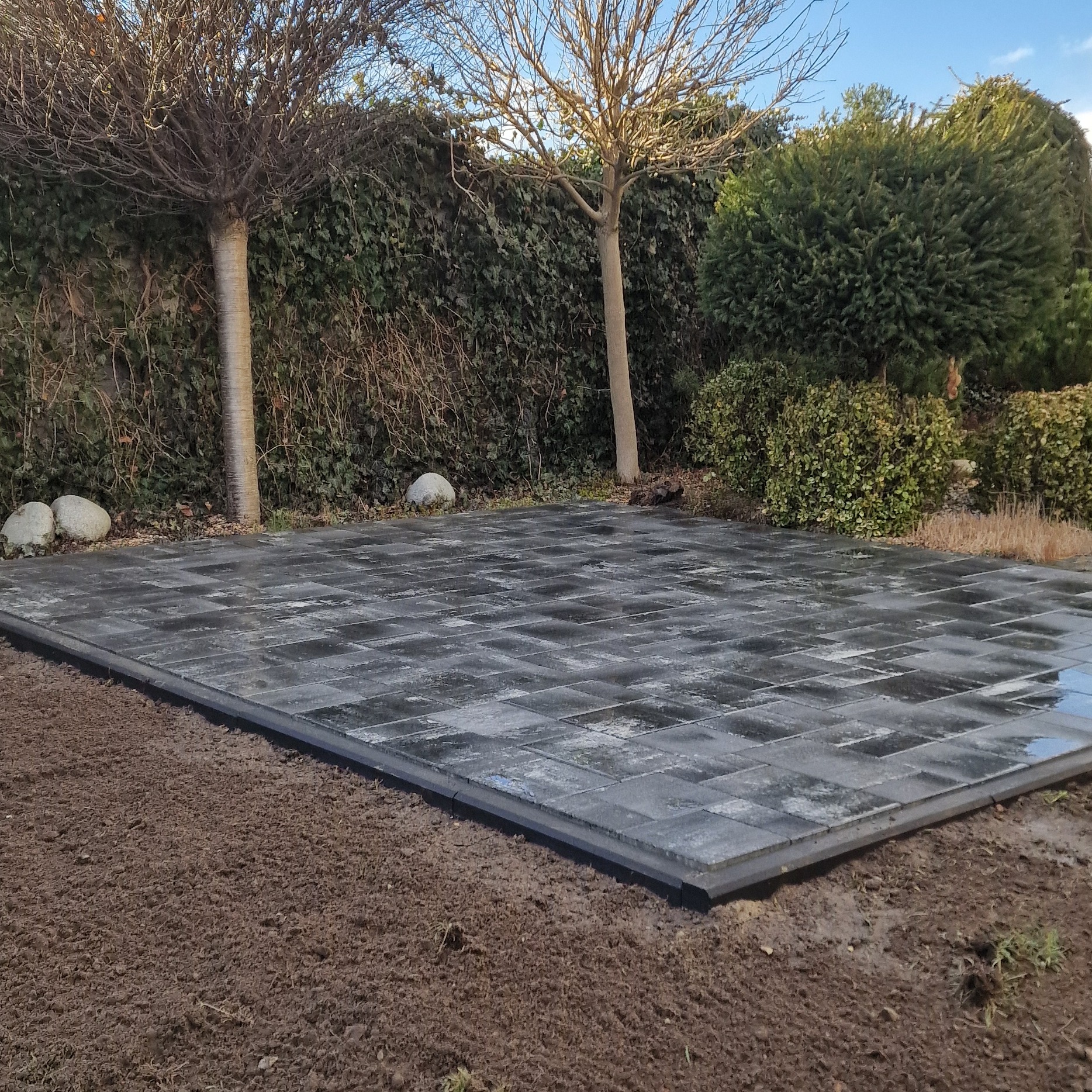 Ułożona szara kostka brukowa na posesji, obrzeża wykończone czarnym elementem. W tle zielony żywopłot, drzewa i fragment błękitnego nieba.