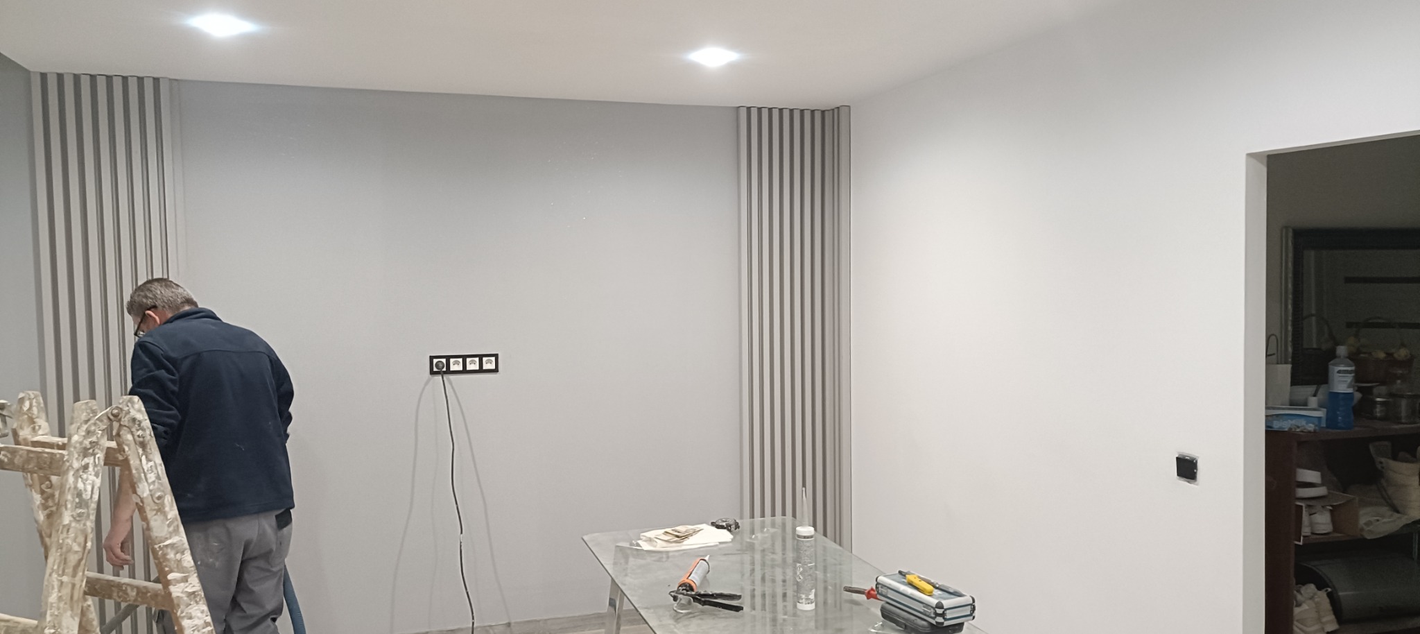 Pracownik na drabinie montuje dekoracyjne panele ścienne w szarym pomieszczeniu z widocznymi gniazdkami elektrycznymi i narzędziami na szklanym stole.