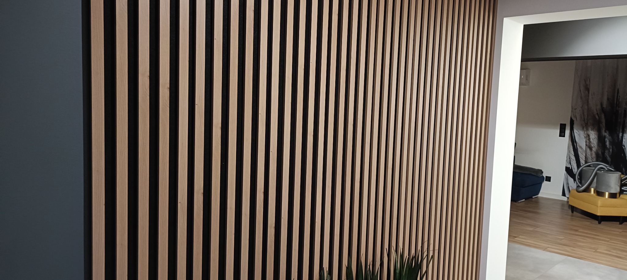Drewniane lamele pionowe na ścianie w kolorze jasnego drewna, widok z boku, w tle widoczne wnętrze pokoju z meblami i dekoracją.