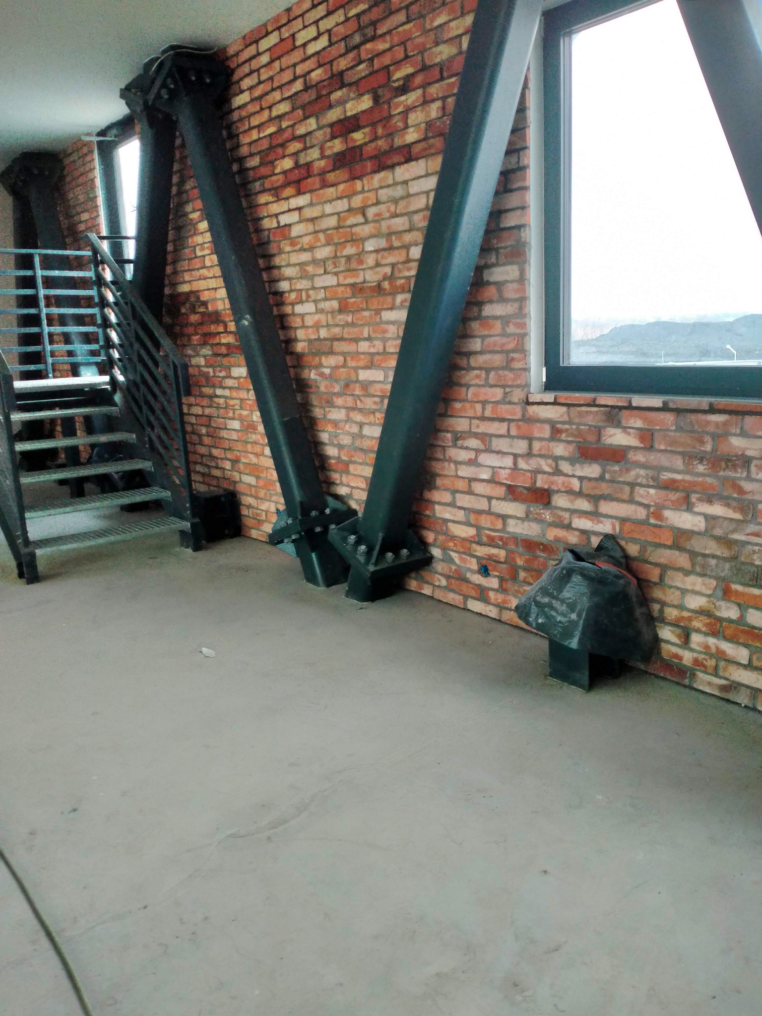 Wnętrze w stylu industrialnym z ceglaną ścianą, metalowymi słupami i schodami, widok na okno z szarym niebem.