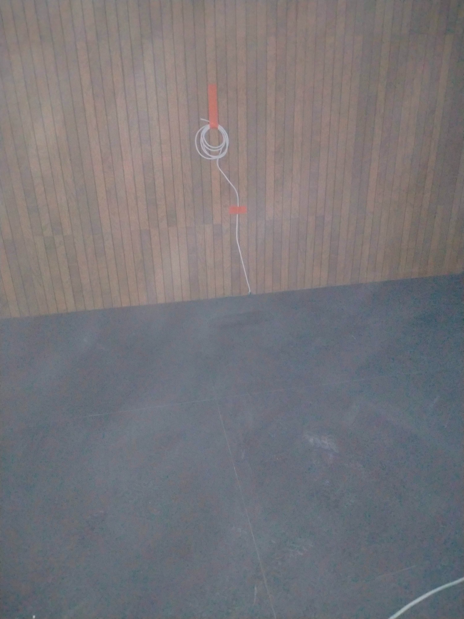Przygotowanie do montażu oświetlenia: kabel zwinięty w pętlę, przymocowany do drewnianej ściany za pomocą pomarańczowej taśmy, widoczny fragment szarej podłogi.
