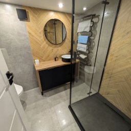 MIKUŚ USŁUGI REMONTOWE I WYKOŃCZENIOWE HYDRAULIKA PIOTR PALAT - Nowoczesna łazienka z kabiną prysznicową typu walk-in, umywalką nablatową na drewnianej szafce z czarnymi frontami i okrągłym lustrem w czarnej ramie, wykończona płytkami imitującymi drewno...