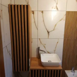 Nowoczesna łazienka z umywalką nablatową, armaturą i szafką z lamelami na tle płytek imitujących marmur.