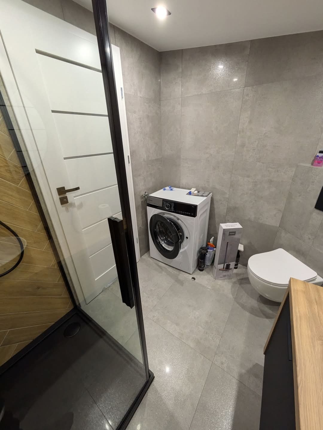 Nowoczesna łazienka z szarymi płytkami, kabiną prysznicową z czarnymi ramami, białą pralką z otwartymi drzwiczkami, toaletą i częścią szafki z drewnianym blatem.