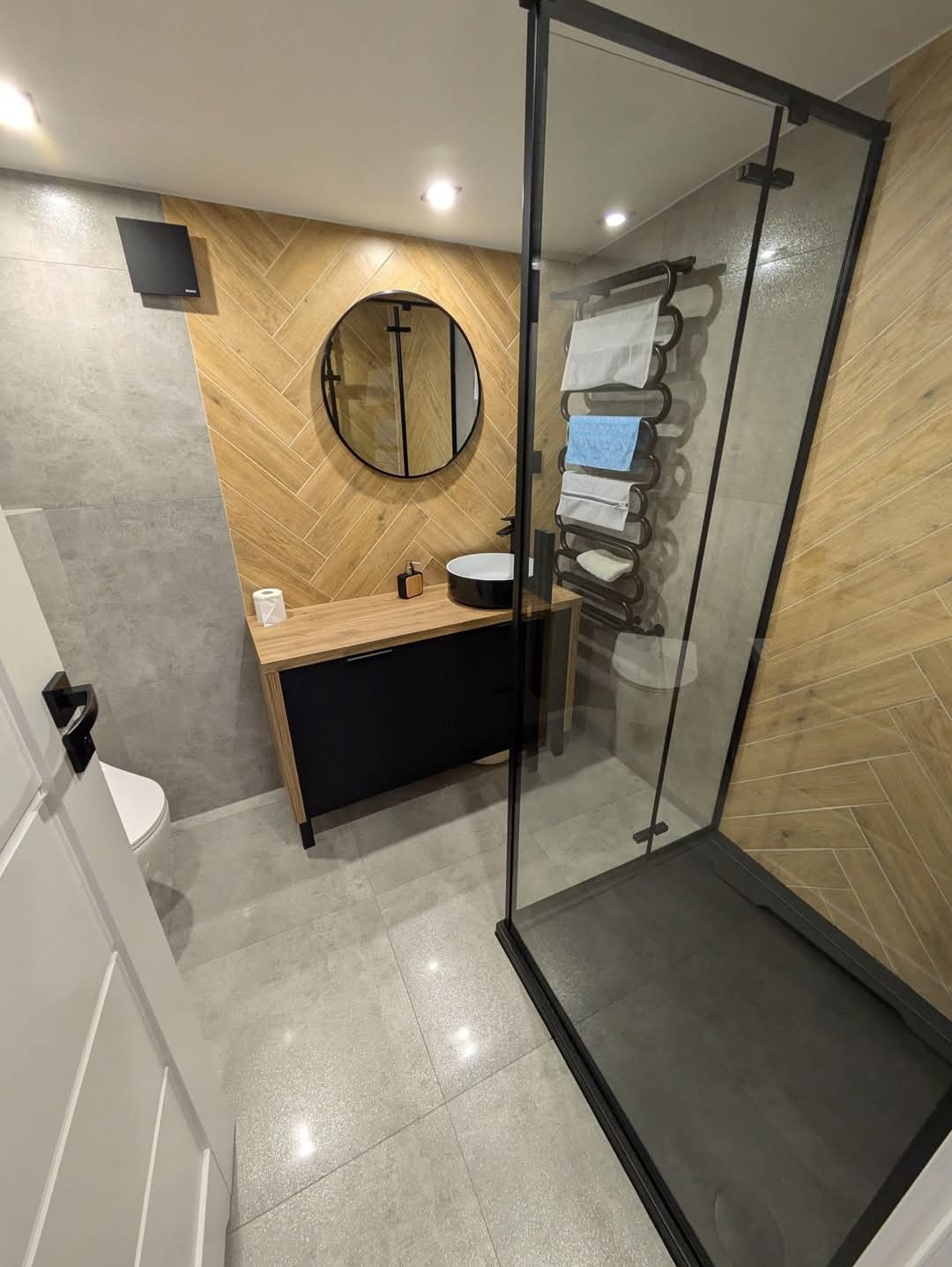 Nowoczesna łazienka z kabiną prysznicową typu walk-in, umywalką nablatową na drewnianej szafce z czarnymi frontami i okrągłym lustrem w czarnej ramie, wykończona płytkami imitującymi drewno...