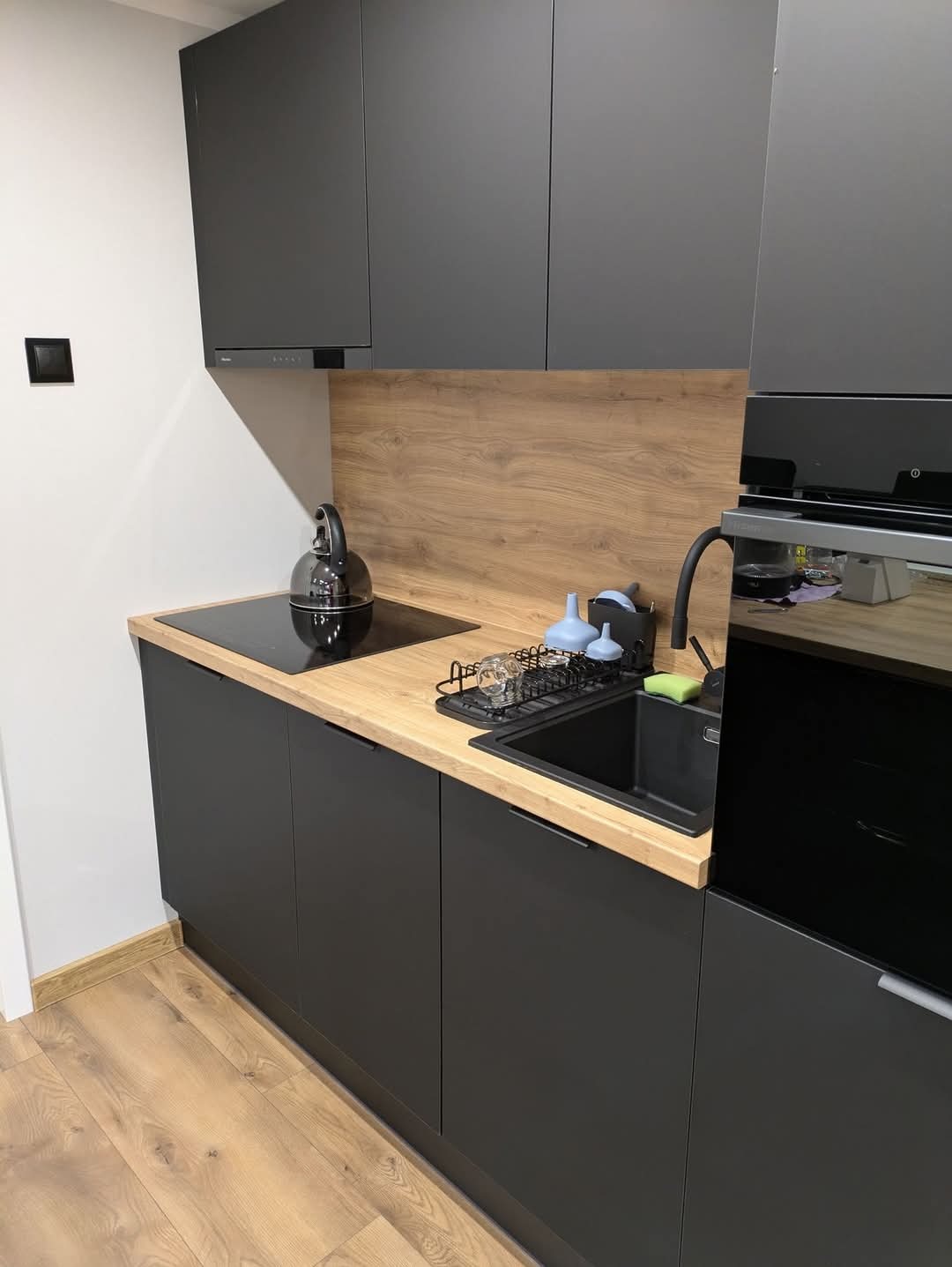 Nowoczesna, minimalistyczna kuchnia z czarnymi matowymi szafkami, drewnianym blatem i panelem ściennym, czarnym zlewem i płytą indukcyjną, czajnikiem na płycie i suszarką do naczyń przy zlewie.