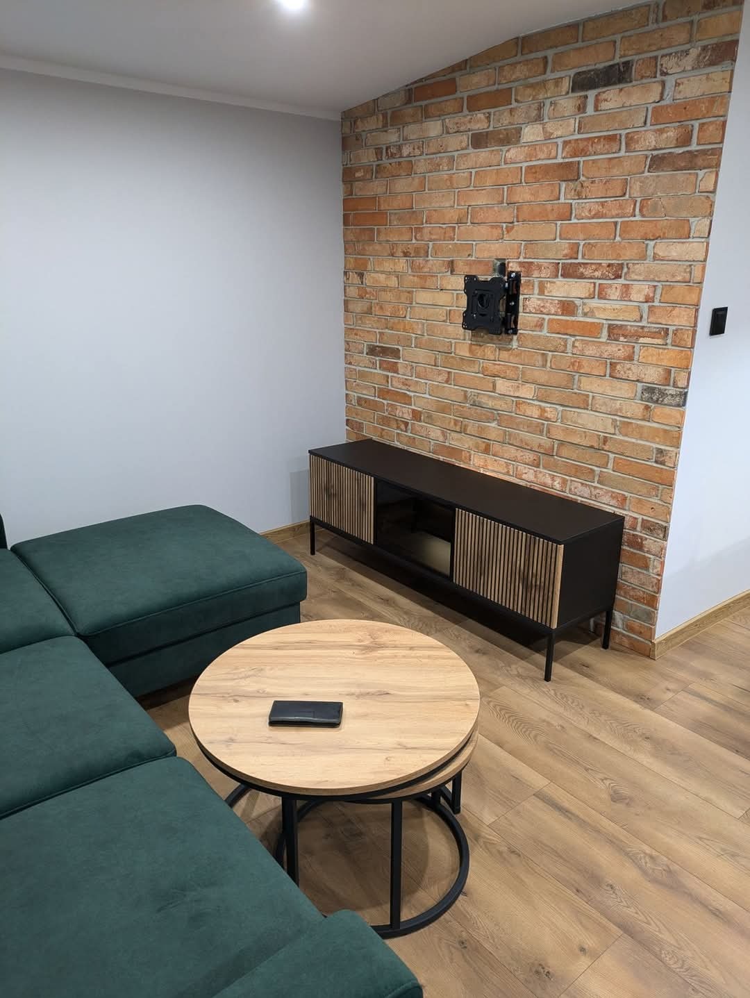 Salon z ceglaną ścianą, ciemnozielonym narożnikiem, okrągłym stolikiem kawowym i czarną szafką RTV z drewnianymi lamelami, na ścianie uchwyt do telewizora.