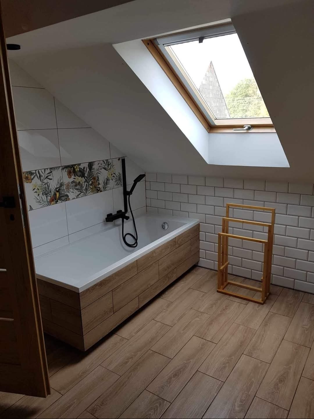 Wykończona łazienka na poddaszu w Strzyżewie z wanną obudowaną drewnopodobną płytką, białą glazurą imitującą cegiełki i oknem dachowym.