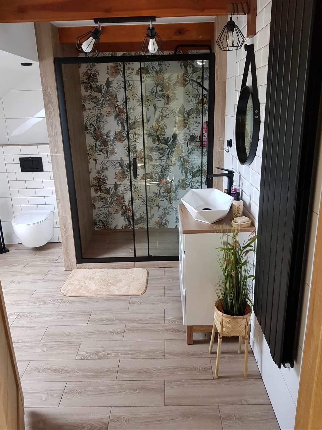 Nowoczesna łazienka z kabiną prysznicową z motywem florystycznym, umywalką nablatową, czarnym grzejnikiem drabinkowym i drewnopodobną podłogą.