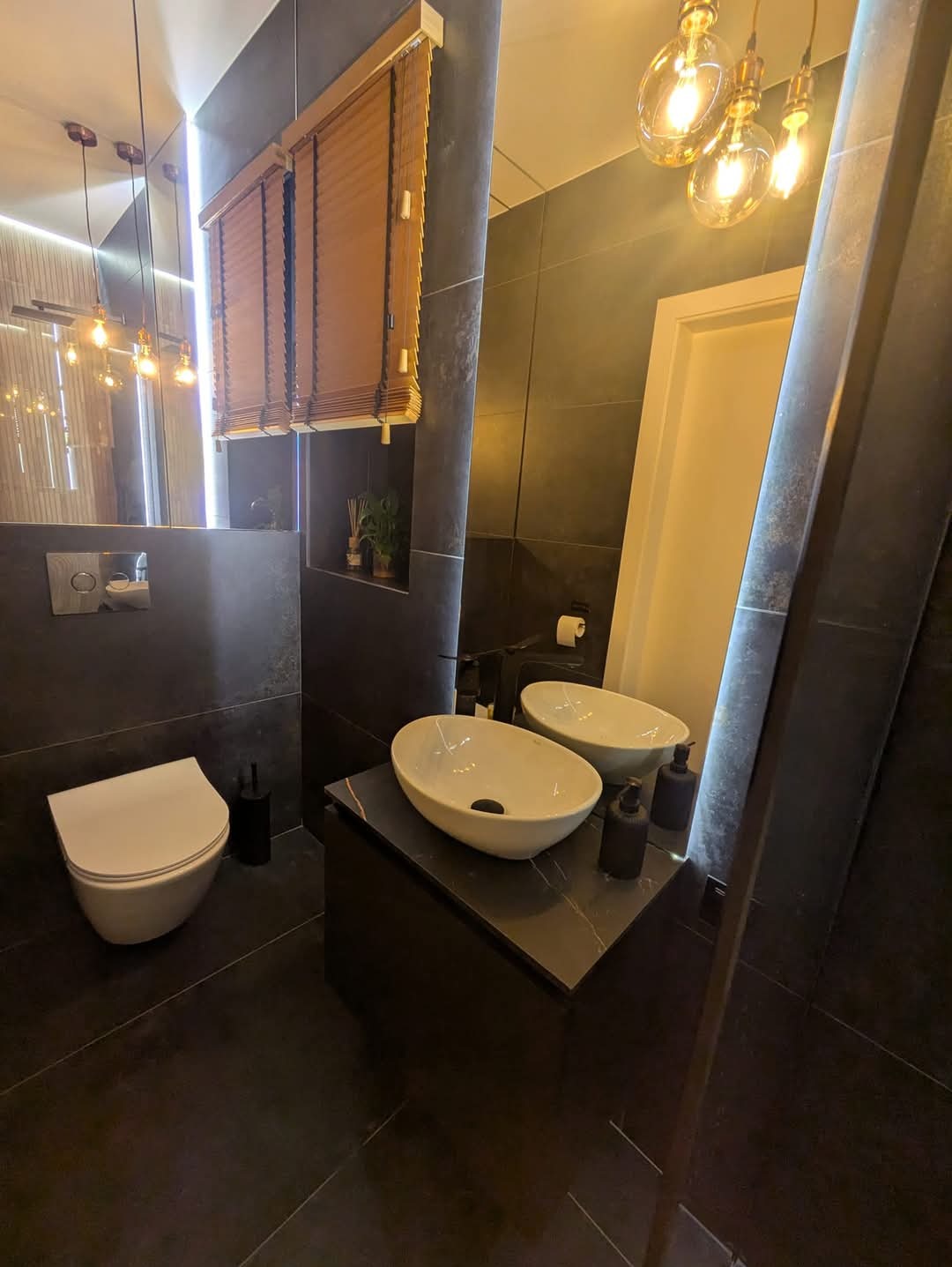 Nowoczesna łazienka z ciemnymi płytkami, podwójną umywalką nablatową, wiszącą toaletą i drewnianymi żaluzjami, oświetlona dekoracyjnymi żarówkami Edison