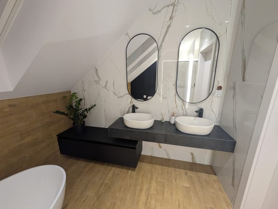 Nowoczesna łazienka na poddaszu z dwoma owalnymi umywalkami nablatowymi, czarną armaturą, podwieszaną szafką i dużymi lustrami w czarnych ramach na tle marmurowych płytek.