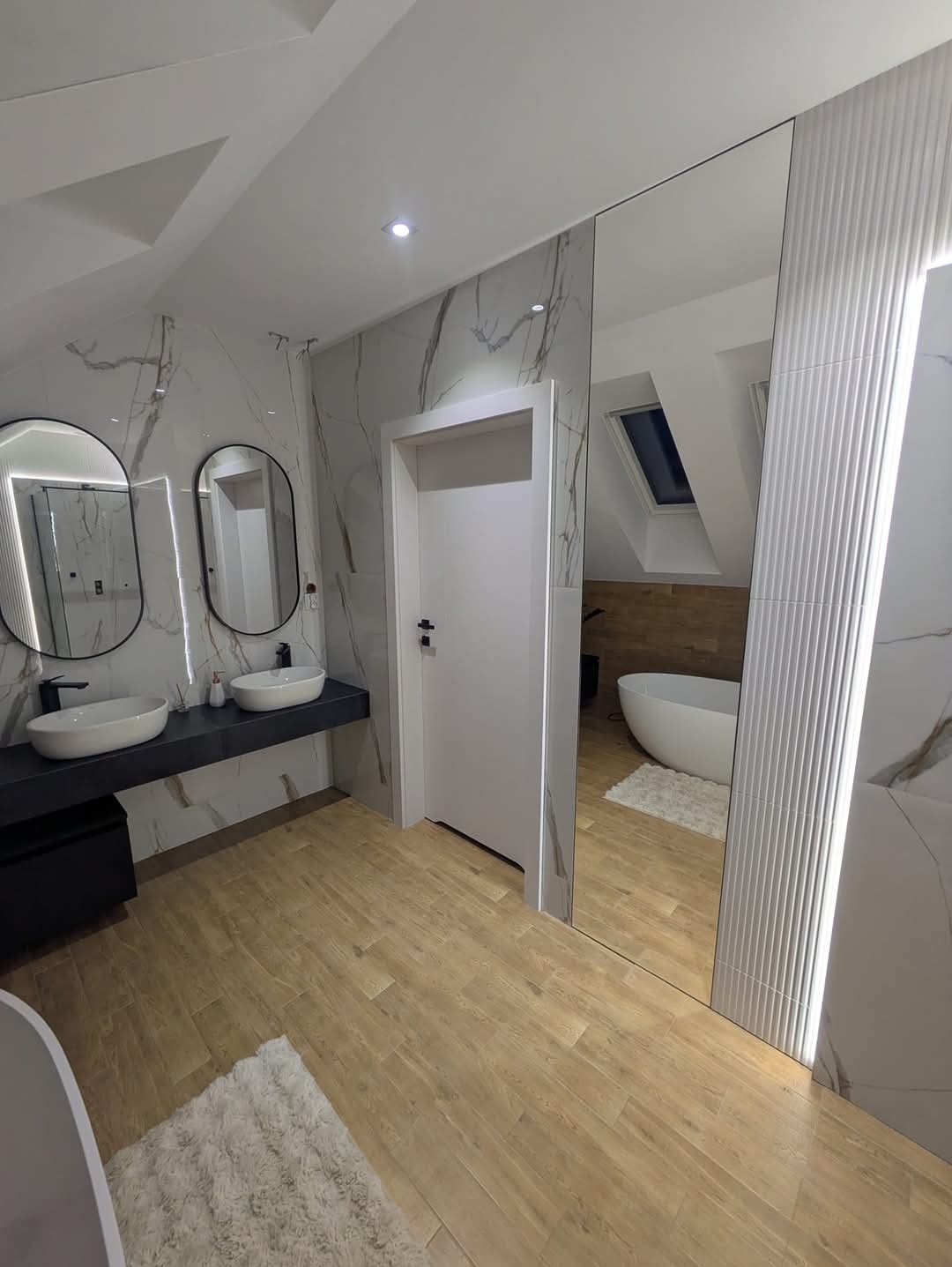 Nowoczesna łazienka na poddaszu z drewnopodobną podłogą, marmurowymi płytkami ściennymi, owalnymi lustrami z podświetleniem LED, białymi umywalkami nablatowymi, czarnym blatem i szafką, białą wanną...