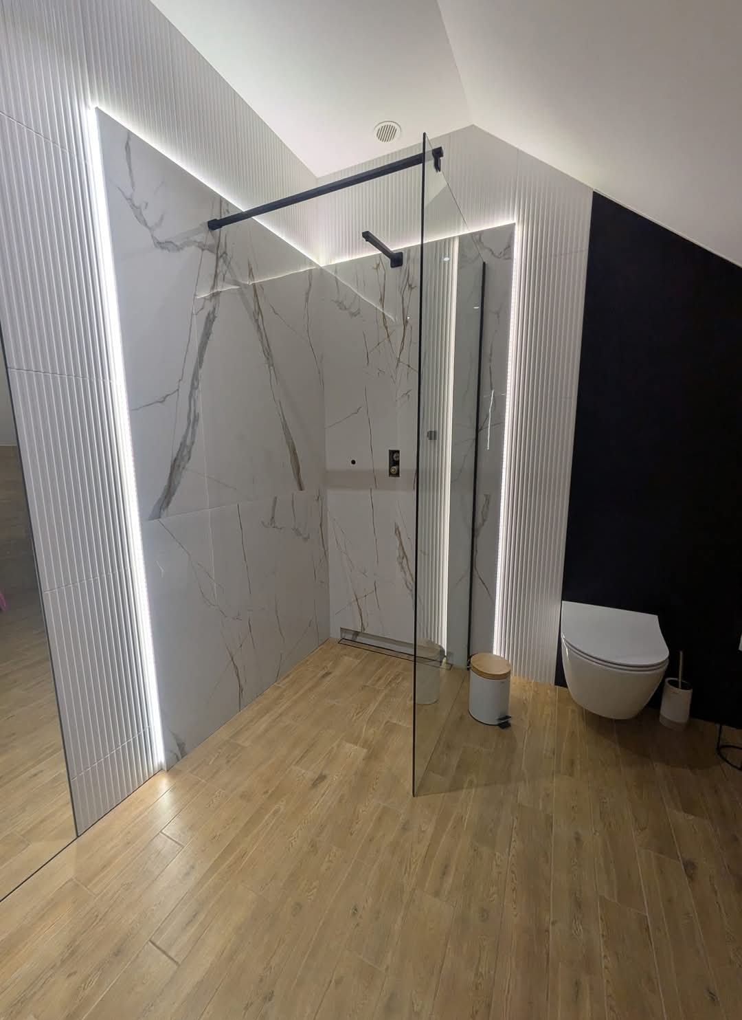 Nowoczesna łazienka z kabiną prysznicową typu walk-in, marmurowymi płytkami na ścianach z subtelnym oświetleniem LED, drewnopodobną podłogą i czarną ścianą z wiszącą toaletą.