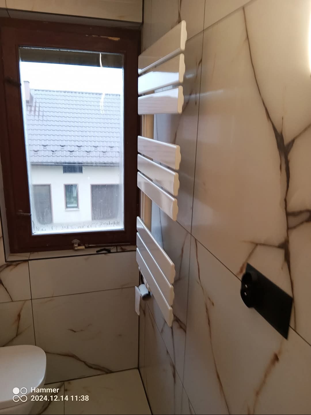 Biały grzejnik łazienkowy drabinkowy zamontowany na ścianie z płytek imitujących marmur, widoczny fragment toalety, okno z widokiem na dom.