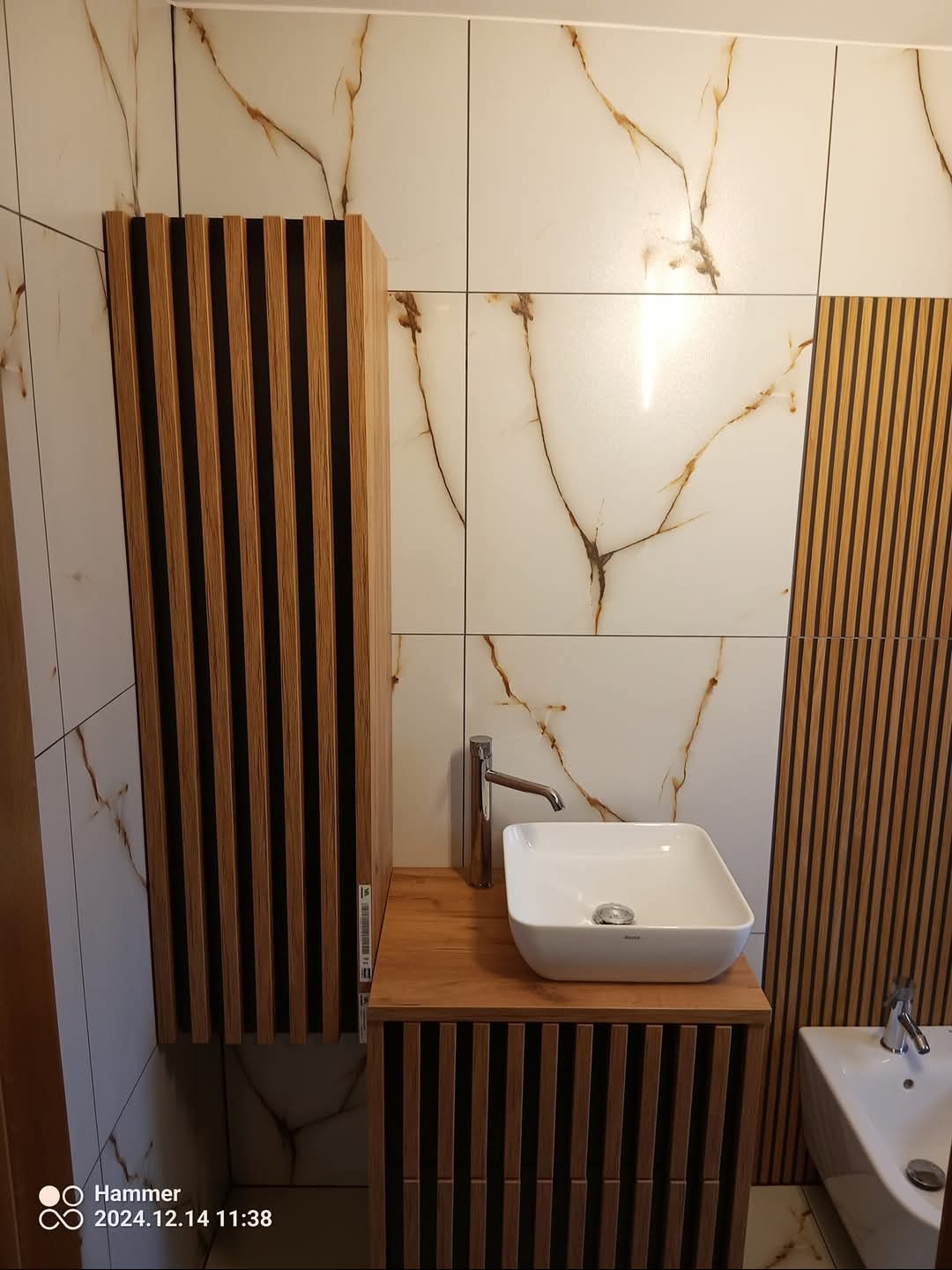 Nowoczesna łazienka z umywalką nablatową, armaturą i szafką z lamelami na tle płytek imitujących marmur.