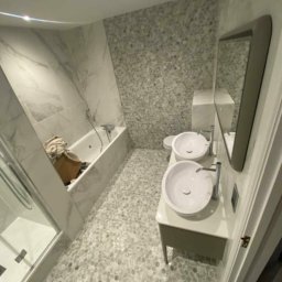 Anna Adamiak - Nowoczesna łazienka z dwoma umywalkami nablatowymi, mozaikową ścianą i podłogą wyłożoną drobnymi kamieniami, widok z perspektywy wejścia.