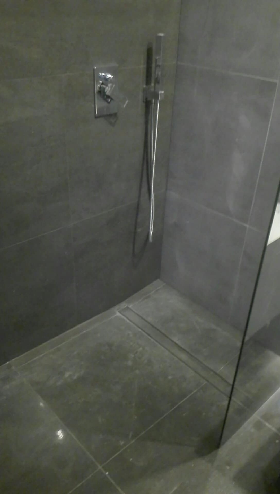 Nowoczesna kabina prysznicowa z ciemnymi, kwadratowymi płytkami, chromowaną armaturą i odpływem liniowym w podłodze.