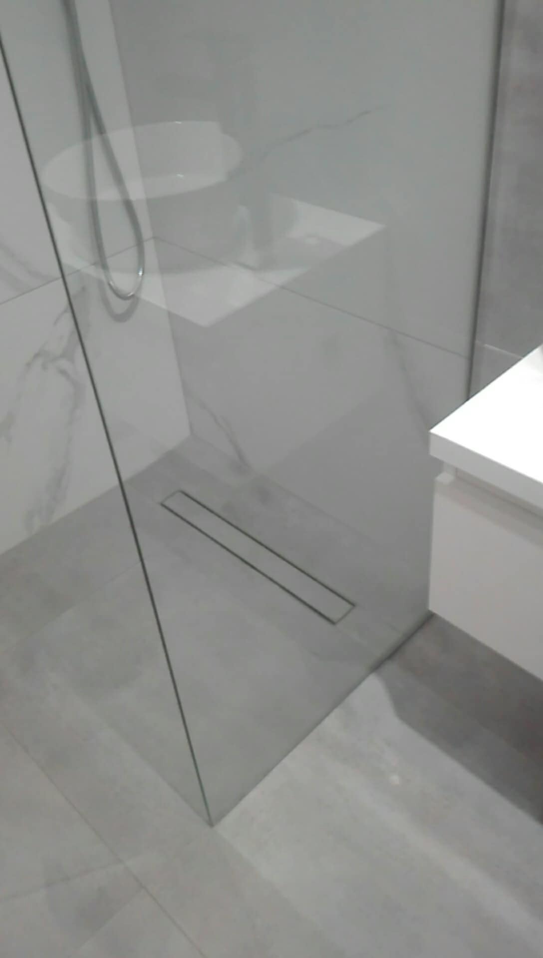 Nowoczesna kabina prysznicowa z odpływem liniowym w posadzce, szklana ściana z widocznym odbiciem umywalki, szare płytki podłogowe i marmurowe ściany.