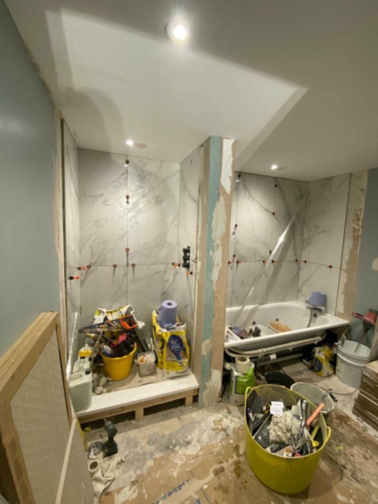 Łazienka w trakcie remontu: białe płytki imitujące marmur na ścianach, krzyżyki dystansowe, przygotowana wanna, widoczne narzędzia i materiały budowlane, surowy stan wykończenia ścian.